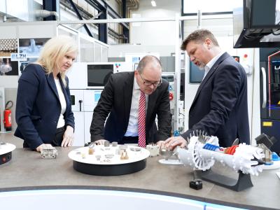 Zwei Männer und eine Frau stehen an einer Fläche mit 3D-Druck-Exponaten und schauen sich diese interessiert an.